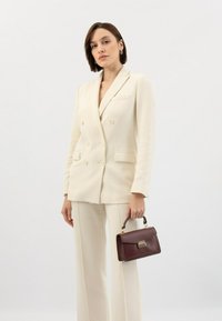 Lichtbeige dubbelzijdige blazer met bijpassende broek; handtas in diep bordeauxleer met gouden hardware en een bovenhandvat.