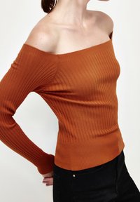Schulterfreier, gerippter Pullover in orange-braun, figurbetontes Design mit langen Ärmeln und strukturierter Oberfläche. Kombiniert mit schwarzen, hochgeschnittenen Hosen.