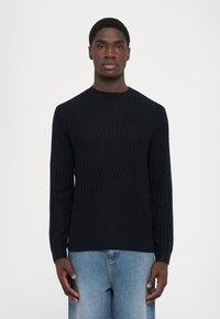 Ung mann iført en marineblå teksturert genser og lyseblå jeans, stående mot en enkel hvit bakgrunn.