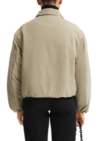 Personne portant une veste beige courte avec un ourlet élastique et un pantalon noir, tenant une chaîne noire dans la main droite, vue de derrière.