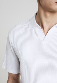 Polo bianco a maniche corte con una texture a coste, dotato di colletto a V e una finitura liscia e pulita.