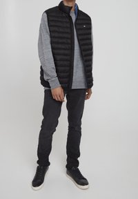 Schwarze Steppweste mit Reißverschluss und hohem Kragen, getragen über einem grauen Sweatshirt. Schwarze Hose und Sneaker vervollständigen das Outfit.