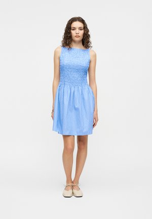 Jonge vrouw met krullend haar staat, gekleed in een lichtblauwe mouwloze jurk met geplooide lijfje en beige platte schoenen met veters tegen een witte achtergrond.