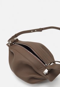 Brown leather bag with a rounded shape, visible zip closure, and a sleek strap featuring adjustable holes and metallic hardware.