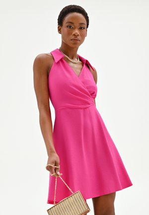 Donna che indossa un vestito rosa senza maniche con dettagli sul colletto, che tiene in mano una borsa a righe dorate, su uno sfondo bianco semplice.