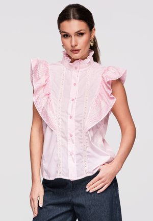 Kvinde iført en ærmeløs lyserød bluse med blondedetaljer og flæseærmer, kombineret med mørke bukser, stående med den ene hånd i lommen.