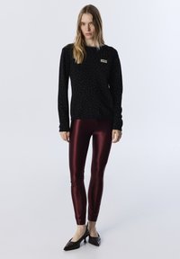 Pull noir avec des pois argentés, col rond. Leggings bordeaux brillants, coupe ajustée. Chaussures à talons noires avec un motif texturé.