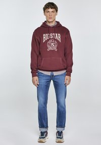 Bordeauxroter Hoodie mit "BIG STAR"-Logo, Kängurutasche, Stofftextur. Kombiniert mit hellblauen Jeans und blauen Sneakers mit beigen Akzenten.