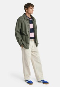 Grön bomulljacka, marinblå och rosa randig polo, beige byxor, blå och vita sneakers, stående pose med händerna i fickorna.