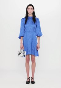 Blauwe gestructureerde jurk met opstaande mouwen, een taille die getailleerd is en een uitlopende rok, gecombineerd met een zilveren handtas en zwarte schoenen met stippen.