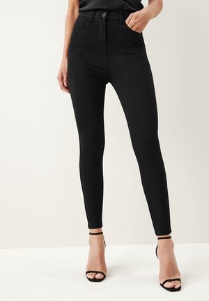 Kobieta ubrana w czarne obcisłe dżinsy z wysokim stanem i czarne sandały na obcasie z paskami wokół kostek, stojąca na tle jasnego, gładkiego tła.