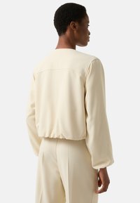 Jasnokremowa bluzka z marszczonym pasem, długimi rękawami i zaokrąglonym dekoltem. Gładka tkanina o subtelnym połysku, minimalne szczegóły szwów.