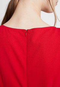 Rode jurk met een gladde textuur, voorzien van een kleine rits aan de achterkant. De halslijn is breed en de stof oogt lichtgewicht.