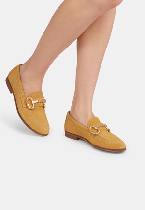 Suede loafers in mustard yellow with a round metal hardware accent, stitched detailing along the edges, and a low brown heel.