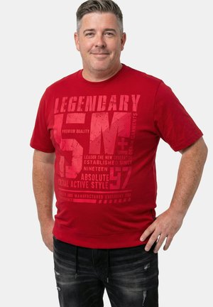 Mann, der mit kurzem Haar lächelt, trägt ein rotes T-Shirt mit Grafik und schwarze zerfetzte Jeans und steht vor einem schlichten weißen Hintergrund.