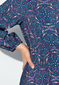 Hand auf der Taille ruhend, gekleidet in einem blauen Kleid mit langen Ärmeln und einem bunten geometrischen Blumenmuster.