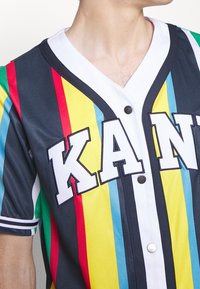 Kleurrijk jersey met korte mouwen, voorzien van verticale strepen in rood, geel, blauw en groen. Met knoopsluiting aan de voorkant, witte kraag en gedurfde "KAN"-letters.