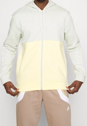 Mann trägt einen zweiteiligen hellgrauen und blassgelben Adidas Kapuzenpullover mit Reißverschluss und beige Adidas Trainingshose und richtet den Saum des Kapuzenpullovers.