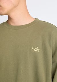 Olívazöld pulóver puha anyagból, bordázott kerek nyakkivágással és világos bézs "nike" logóval az mellkason.