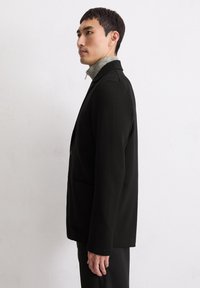 Schwarzer Blazer mit einem klaren Design,Ein-Knopf-Verschluss und Reverskragen. Getragen über einem grauen hochgeschlossenen Oberteil, das eine glatte Stofftextur aufweist.