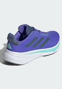 Zapatilla deportiva morada con parte superior de malla transpirable, detalles en gris y una entresuela blanca que cuenta con una franja verde menta. Se ve el logo de las tres franjas.