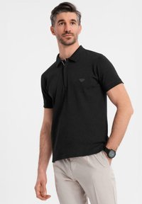 Černé polo tričko s dvoupřezkovým límečkem, krátkými rukávy a malým trojúhelníkovým logem na levém prsou. Nosené s světlými kalhotami.