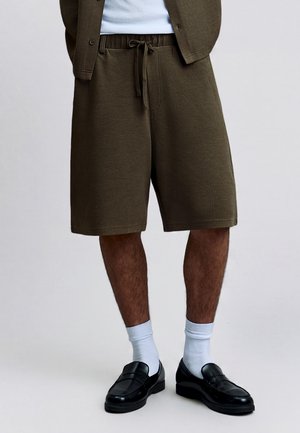 Mand iført teksturerede olivengrønne shorts til knæet med snøre, hvide sokker og sorte læderloafers, stående mod en ensfarvet baggrund.