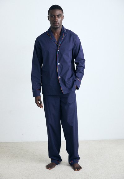 Set di pigiami in cotone blu navy con camicia abbottonata e rifiniture arcobaleno e pantaloni coordinati, indossati da un modello a piedi nudi su uno sfondo neutro.
