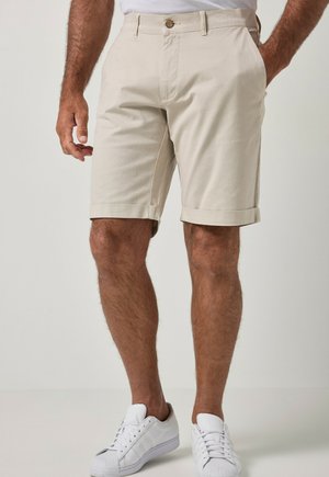 Mand iført beige shorts med rullet kant, knap og lynlås, hvide sneakers og en hvid skjorte, der er stoppet ned i bukserne, stående med den ene hånd i lommen.