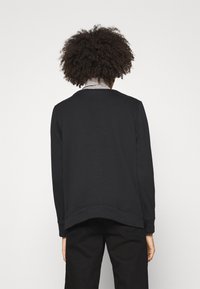 Sudadera negra con puños y dobladillo de canalé, que presenta un corte relajado. Llevada sobre una camisa a rayas, mostrando una textura suave.