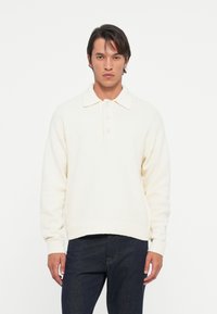 Pulover de culoare crem, cu guler polo, trei butoane, manșete și tiv cu nervuri. Material texturat moale; croială lejeră.