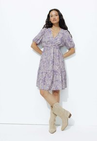 Robe à imprimé floral violet avec des manches bouffantes, un décolleté en V et une jupe à volants, assortie à des bottines en daim tan. Texture lisse.