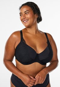 Reggiseno nero con dettagli in pizzo e coppe lisce. Spalline regolabili e supporto con ferretto. Indossato da una modella, in evidenza su uno sfondo neutro.