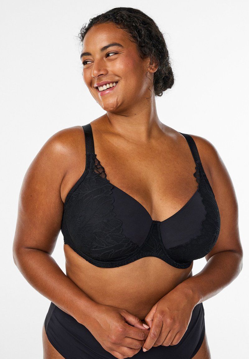 Reggiseno nero con dettagli in pizzo e coppe lisce. Spalline regolabili e supporto con ferretto. Indossato da una modella, in evidenza su uno sfondo neutro.