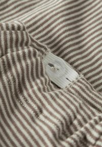 Gestreepte stof in beige en bruin, met een gladde textuur, voorzien van een zichtbare witte knop en sluiting. Focus op de stikseldetails.