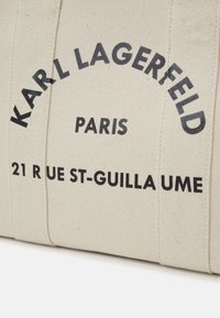 Beige canvas tas met zwarte bedrukte tekst: "KARL LAGERFELD PARIS 21 RUE ST-GUILLAUME," met gestikte details langs de zijkanten.