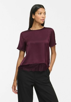 Femme portant un chemisier violet en soie à manches courtes avec un bord en dentelle à l'ourlet et un pantalon noir, regardant sur le côté avec une main dans la poche.