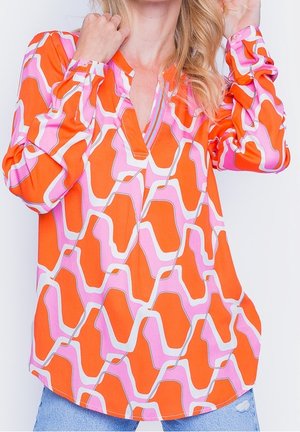 Woman wearing a long-sleeve orange, pink, and white geometric patterned blouse with V-neckline and blue distressed jeans.