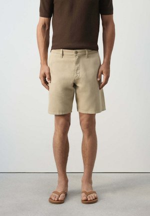 Hombre vistiendo pantalones cortos beige, camisa marrón de manga corta y sandalias marrones, de pie contra un fondo claro y liso.