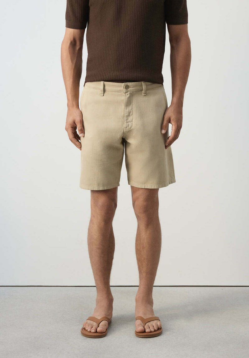 Homme portant un short beige, une chemise marron à manches courtes et des sandales marron, debout devant un fond clair uni.