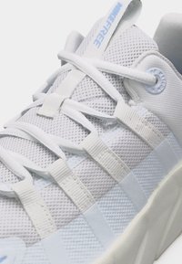 Κοντινό πλάνο σε λευκό αθλητικό παπούτσι με ύφασμα πλέγματος, παχιά σόλα και κορδόνια περασμένα μέσα από υφασμάτινες θηλιές, με το κείμενο "NIKE FREE" στη γλώσσα.