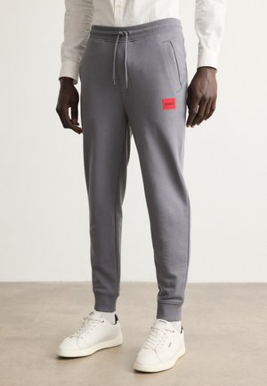 Pantaloni tuta grigi realizzati in misto cotone, dotati di una toppa etichettata rossa e tasche laterali, abbinati a sneakers bianche. Texture liscia, vestibilità affusolata.