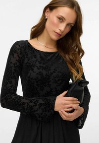 Melna garā piedurkne kleita ar teksturētu ziedu rakstu, apaļu izgriezumu un saplacinātām piedurknēm. Modelis tur nelielu melnu somiņu. Redzama sudraba kaklarota.