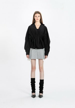 Joven mujer con blusa negra de gran tamaño, falda gris plisada, calentadores de piernas negros y tacones negros puntiagudos, de pie frente a un fondo blanco.