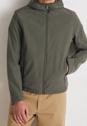Uomo che indossa una giacca con cappuccio verde oliva con cerniera e mani nelle tasche, pantaloni beige, su sfondo chiaro e uniforme.