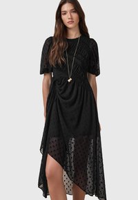 Vestido negro con tejido texturizado, mangas cortas, diseño fruncido, dobladillo asimétrico y sección inferior transparente. Acentuado con un collar de colgante.