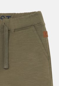 Des joggings vert olive présentent une taille élastique avec cordon de serrage, une poche latérale et une petite étiquette en cuir sur le côté droit. Texture de tissu lisse visible.