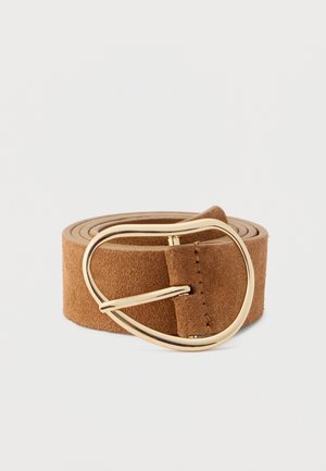 Ceinture en suède marron roulée avec une grande boucle métallique ovale irrégulière dorée sur un fond clair uni.