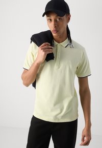 Svetložltý polo tričko s golierom, trojitou gombíkovou légiou a námorníckymi akcentmi. Model drží čierny sveter a nosí čierny klobúk.