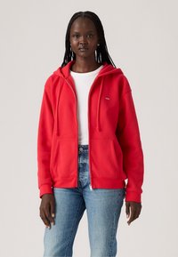 Sweat à zip rouge en tissu doux, avec capuche à cordon, poches avant et un petit logo sur la poitrine gauche. Assorti à un jean clair.
